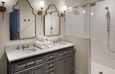 This transitional gray and white master bath remodel includes a large walk-in shower with glass hinged door and white subway tile walls, a double sink vanity paired with gray shaker cabinets and white marble countertops and a matte white hexagon floor tile. The brass sconces and vintage arched mirrors complete the look of this master bath remodel.