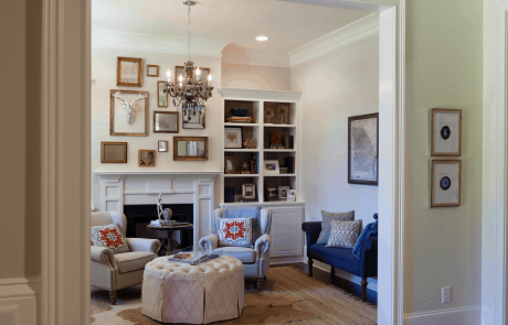 This large traditional foyer and front room features dark brown hardwood floors, beige walls, a stunning white fireplace, and custom built in bookcases. The blue velvet bench, wingback chairs, round peach tufted ottoman and gold accents add elegance and warmth to the space.