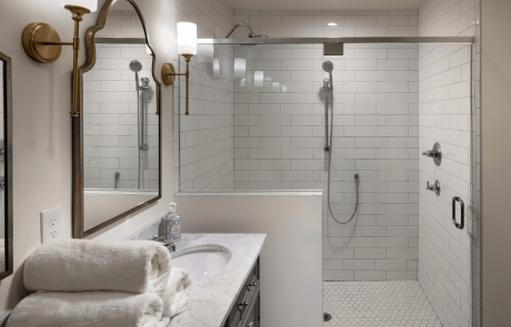 This master bath remodel features a large curbless shower with a white porcelain mosaic floor tile, white subway tile shower walls and a rain shower head with separate handheld spray. A double sink vanity with white marble countertops and dark gray shaker cabinets are accented with antique brass wall sconces and arched mirrors.