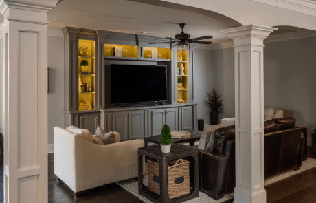 Custom basement remodel with separate TV area features a gray custom built-in entertainment center complete with inset lighting and storage, dark brown hardwood floors and comfortable seating.