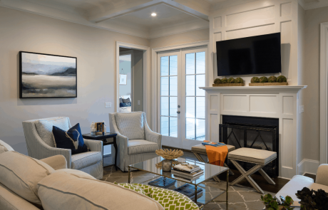 Neutral living room with gray walls, white trim, and medium brown hardwood floors. A flat screen television is mounted to a custom white wainscot fireplace and mantel, flanked by double French doors that lead to a covered outdoor living space for indoor outdoor living. Navy, green and orange accent pillows, and a glass-top metal coffee table add a modern touch and a pop of color to this transitional living room.