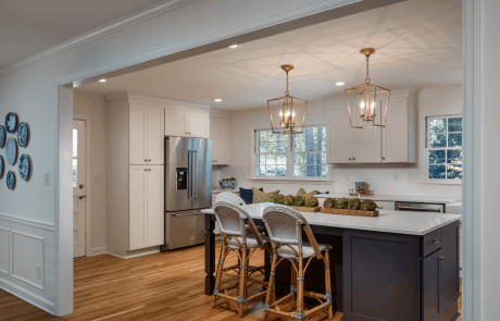 Custom kitchen remodel features a modern farmhouse style kitchen with two toned cabinetry in blue and white. The large island features dark blue cabinets with white quartz countertops and seating for 3. White recessed panel cabinets, and a custom-built vent hood and built-in bench give the room a fresh and modern look.