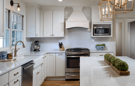 Modern farmhouse kitchen with large island, white quartz countertops, white recessed panel cabinets and custom vent hood, white farmhouse sink, white apron sink, sleek white subway tile backsplash and stainless-steel appliances.