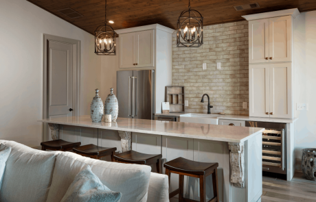 Detached custom pool house addition decorated in neutral tones features a vaulted cedar tongue & groove ceiling and full custom kitchen with bar island, white quartz countertops, soft white cabinetry, and a sand-colored matte porcelain subway tile backsplash. A set of modern dark bronze chandeliers and decorative antiqued corbels add a hint of luxury to the space.