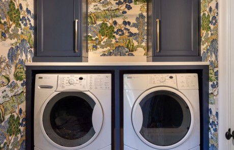 Blue, green & white laundry room addition with side-by-side washer & dryer, blue custom shaker cabinets, bronze hardware, white hexagon porcelain mosaic floor, a hanging drying station, hidden ironing board and broom closet.