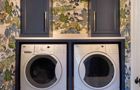 Blue, green & white laundry room addition with side-by-side washer & dryer, blue custom shaker cabinets, bronze hardware, white hexagon porcelain mosaic floor, a hanging drying station, hidden ironing board and broom closet.