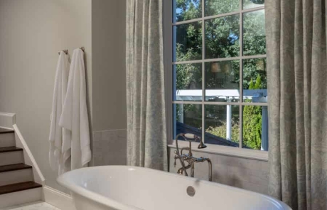 Free standing white soaking tub with polished nickel freestanding tub filler, white marble floor, gray walls, and silver semi flush mount ceiling light.