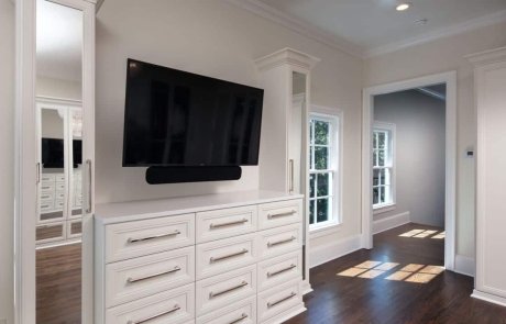 Second story addition with large private dressing room, white custom cabinetry with polished nickel hardware and mounted tv, brown hardwood flooring, and walls painted in Dove Wing by Benjamin Moore.