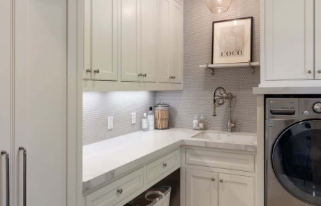 Custom Laundry Room with gray porcelain tile floor and penny round grey porcelain tile backsplash, white custom cabinets with floor to ceiling storage, solid surface countertops and undermount sink with Waterstone faucet.
