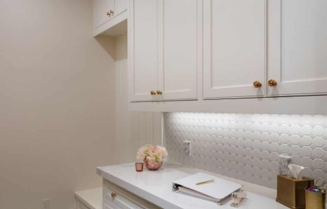 Mudroom remodel has a gray and white diamond pattern tile floor, white custom cabinets and built-ins with aged brass hardware, porcelain countertops and a matte white octagon mosaic backsplash.
