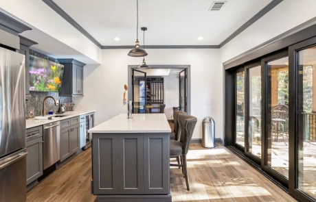 Open concept basement renovation gets plenty of natural light with black panoramic sliding doors providing an instant indoor-outdoor connection to the outdoor living space. A freestanding bar with gray shaker cabinetry and white quartz cabinets is home to a built-in kegerator while a fully kitchen provides all the necessities for entertaining.