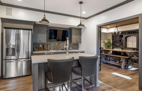 This large basement remodel features a fully equipped contemporary shaker kitchen and bar with grey shaker cabinets, gray and white glass mosaic backsplash, white Viatera Minuet quartz countertops, stainless steel appliances including a built-in kegerator and light brown porcelain plank tile flooring.