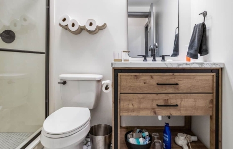 The full bathroom features a freestanding modern rustic vanity with single sink, white and gray quartz countertops, black hardware, a frameless glass shower with white hexagon mosaic tile and a gray matte ceramic tile floor.