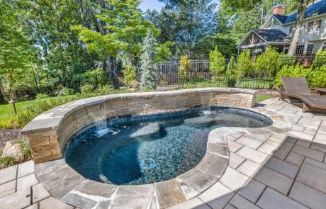 Custom pool design includes a heated plunge pool with sheer descents, flagstone coping, and stacked stone seating wall with a Black Marble PebbleTec finish. Other features include paver patio and backyard pool landscape design.