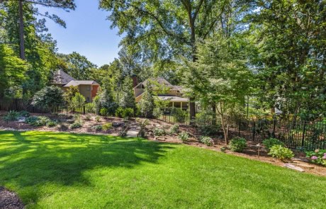 This custom pool and backyard landscape design features a beautiful, landscaped mix of plants, shrubs, and flowers with a lush green lawn. Outdoor lighting creates a stunning backdrop in this revamped outdoor living area when the sun goes down.