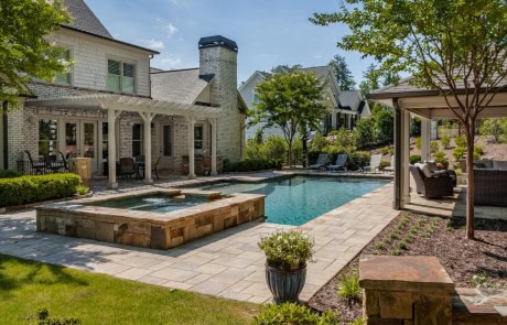 Stunning backyard transformation in Milton, GA offers year-round outdoor living and pool entertainment with features including a rectangular pool with a raised spa, water feature, and tanning ledge, a covered poolside cabana with stacked stone fireplace, pergola, paver patio and thoughtful landscape design.