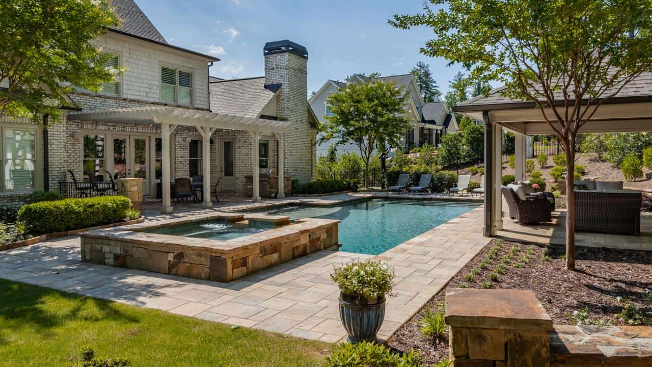 Stunning backyard transformation in Milton, GA offers year-round outdoor living and pool entertainment with features including a rectangular pool with a raised spa, water feature, and tanning ledge, a covered poolside cabana with stacked stone fireplace, pergola, paver patio and thoughtful landscape design.