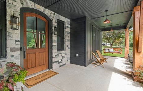 Inviting front porch on a new home build, featuring stone accents, a craftsman-style wood door with divided glass panes, black siding, a beadboard ceiling, and cozy seating.