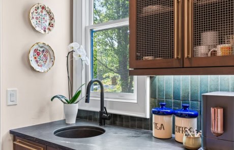 Charming coffee bar with dark wood cabinetry, black soapstone countertop, green tile backsplash, brass hardware, and mesh cabinet doors.