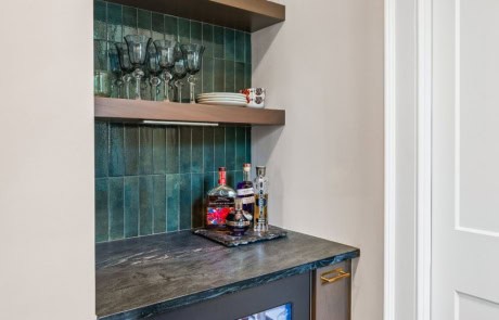 Stylish beverage nook with green tile backsplash, open wood shelves for glassware display, black soapstone countertop, and a built-in beverage fridge with brass hardware.