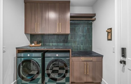 Modern laundry room features a functional design with dark wood cabinetry, black countertops, green tile backsplash, brass hardware, and a checkerboard tile floor.