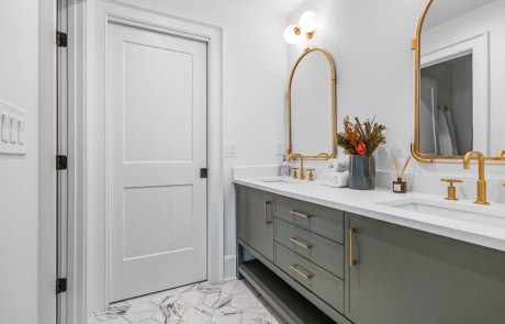 Elegant guest bathroom featuring a double vanity with soft green cabinetry, white quartz countertop, brass fixtures, arched mirrors, and marble-patterned floor tile for a luxurious touch.