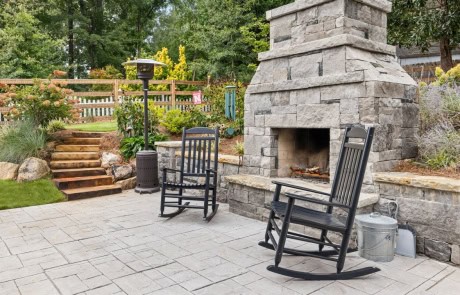 Cozy outdoor patio space featuring a stone fireplace with a seating area, rocking chairs, and a paver patio, surrounded by landscaped garden beds and natural stone steps leading to the backyard.